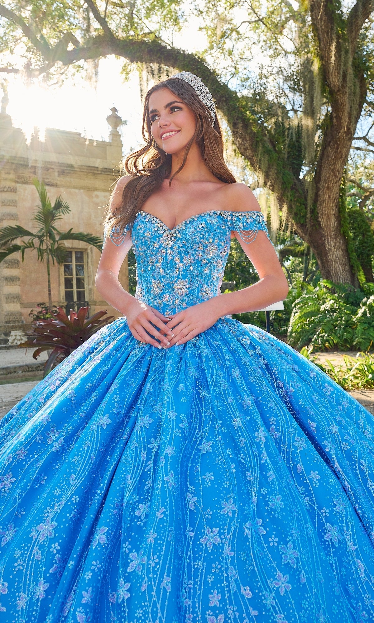 A woman outdoors wears the Quinceanera Dress 54220 By Amarra, a sparkly blue off-the-shoulder ball gown with a floral glitter tulle skirt and decorative headpiece; trees and a building are visible in the background.